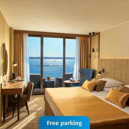 Grand Hotel Bernardin Portorose Esterno foto The photo shows a modern hotel room featuring a large window with a view of the water. The room is decorated in warm tones, with a comfortable bed that has gold-colored bedding. There is a desk and chair situated next to the window, as well as a wall