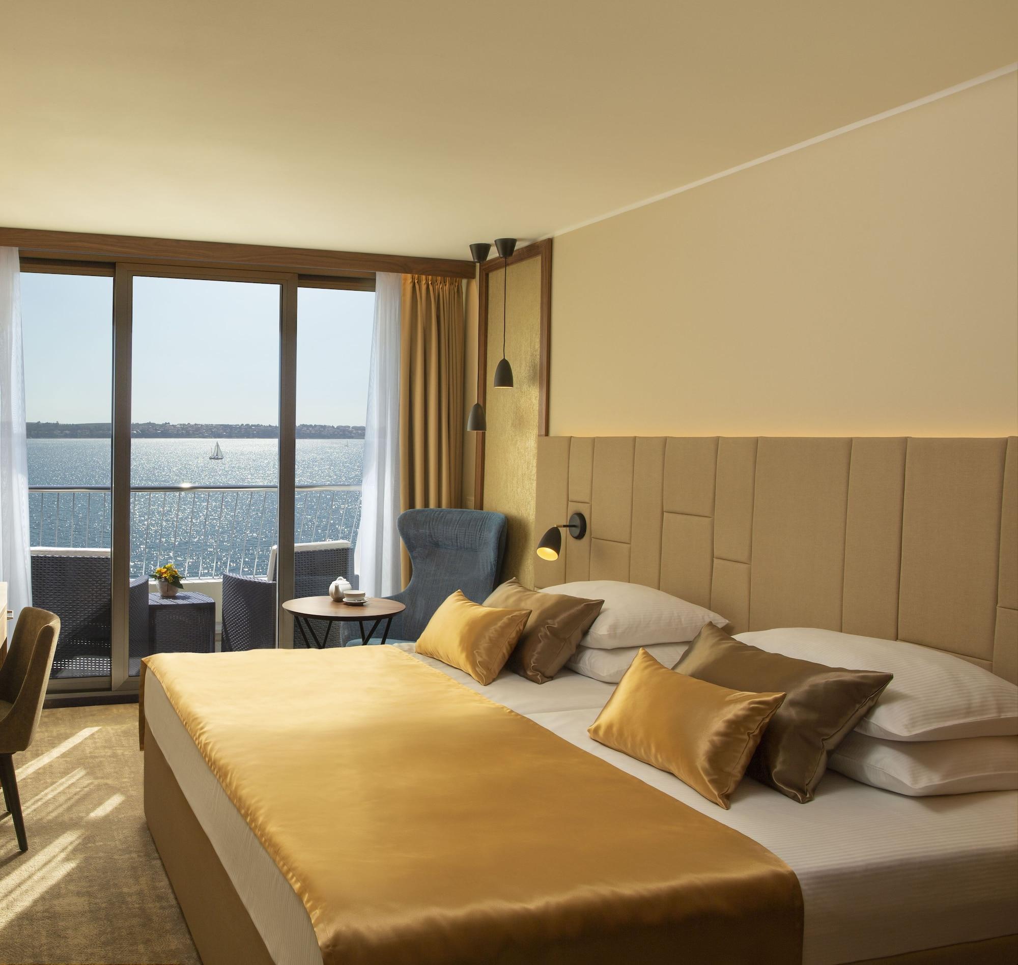 Grand Hotel Bernardin Portorose Esterno foto The photo shows a cozy hotel room with a modern design. The room features a large bed with a beige and gold color scheme, complemented by decorative pillows. There is a seating area with a blue armchair and a small round table, positioned near a larg