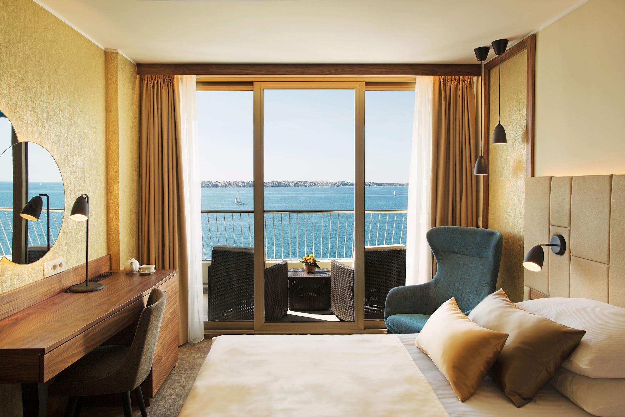 Grand Hotel Bernardin Portorose Esterno foto The image shows a well-designed hotel room with a modern aesthetic. The room features a large bed with decorative pillows and a soft blanket. There's a stylish armchair in a blue hue next to a small table with two chairs, creating a cozy seating area