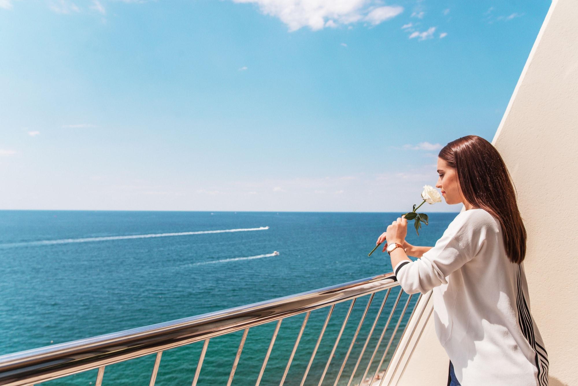 Grand Hotel Bernardin Portorose Esterno foto The photo depicts a woman standing on a balcony overlooking the ocean. She holds a white rose and looks thoughtfully toward the water, which appears calm and blue. There are two boats visible on the surface of the sea, creating gentle waves. The sky 