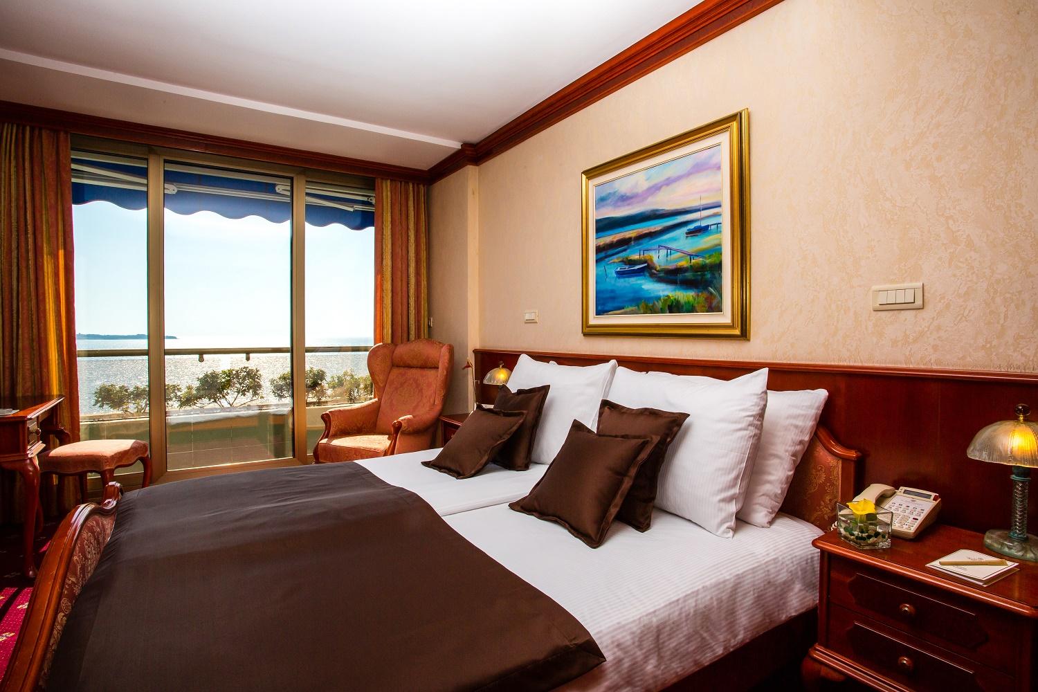 Grand Hotel Bernardin Portorose Esterno foto The photo shows a cozy hotel room featuring a double bed with white bedding and dark brown accents, including decorative pillows. There are two armchairs in the corner, and a large window with a blue awning that provides a view of the outdoors, likel