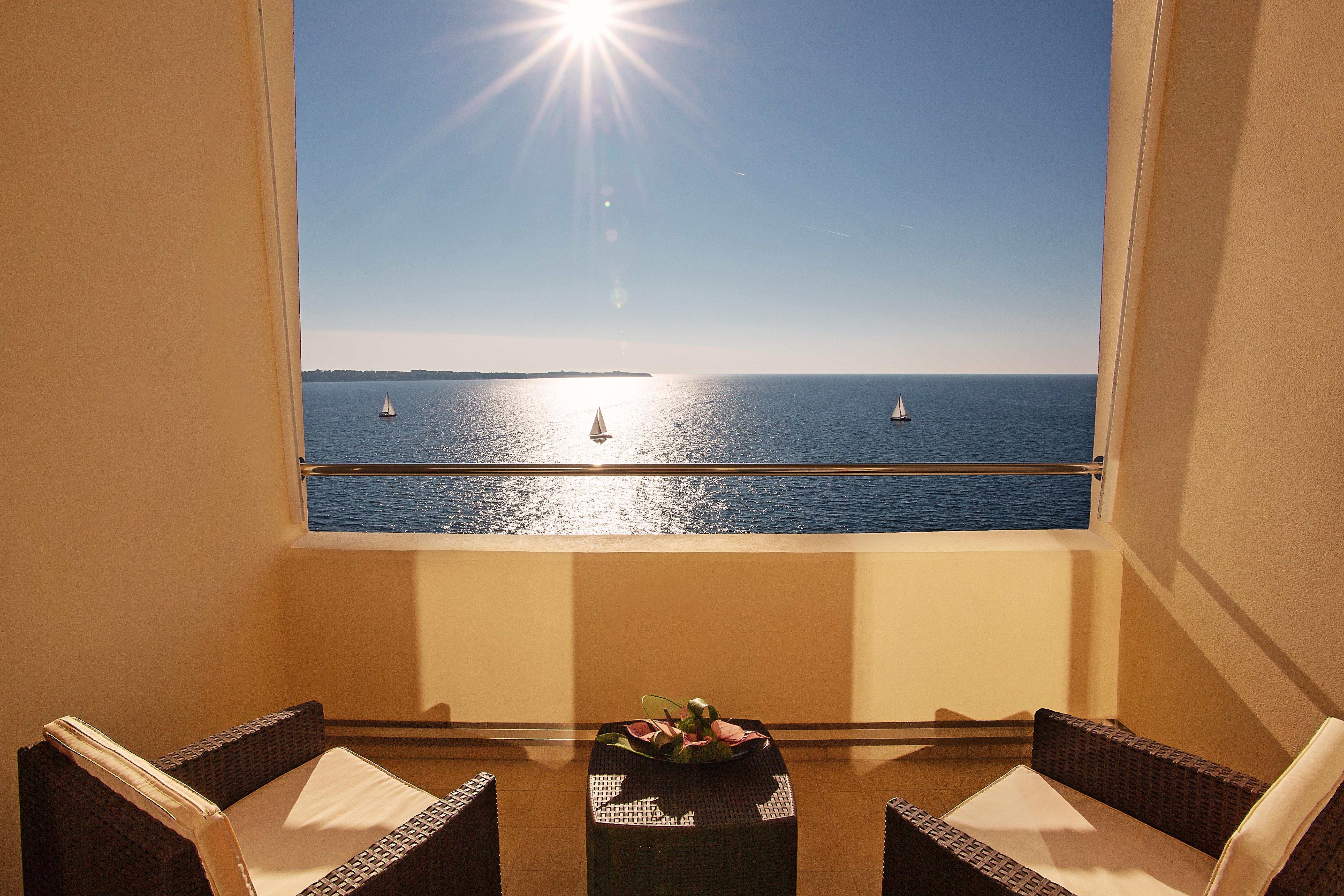 Grand Hotel Bernardin Portorose Esterno foto The photo depicts a serene coastal view from a room or balcony. In the foreground, there are two wicker chairs and a small table, creating a cozy seating area. Beyond the railing, a bright blue sea stretches out under a clear sky, with a shining sun 