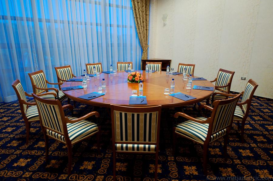 Grand Hotel Bernardin Portorose Esterno foto The photo shows a conference room with a large round wooden table at its center. There are several wooden chairs arranged around the table, each with striped upholstery. On the table, there are folded blue napkins, water bottles, and a small floral c