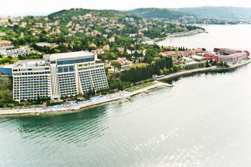 Grand Hotel Bernardin Portorose Esterno foto The image shows an aerial view of a waterfront area. There is a large modern hotel or building with a lot of windows situated near the shore. Alongside the building, there is a beach area with what appears to be sun loungers. In the background, there