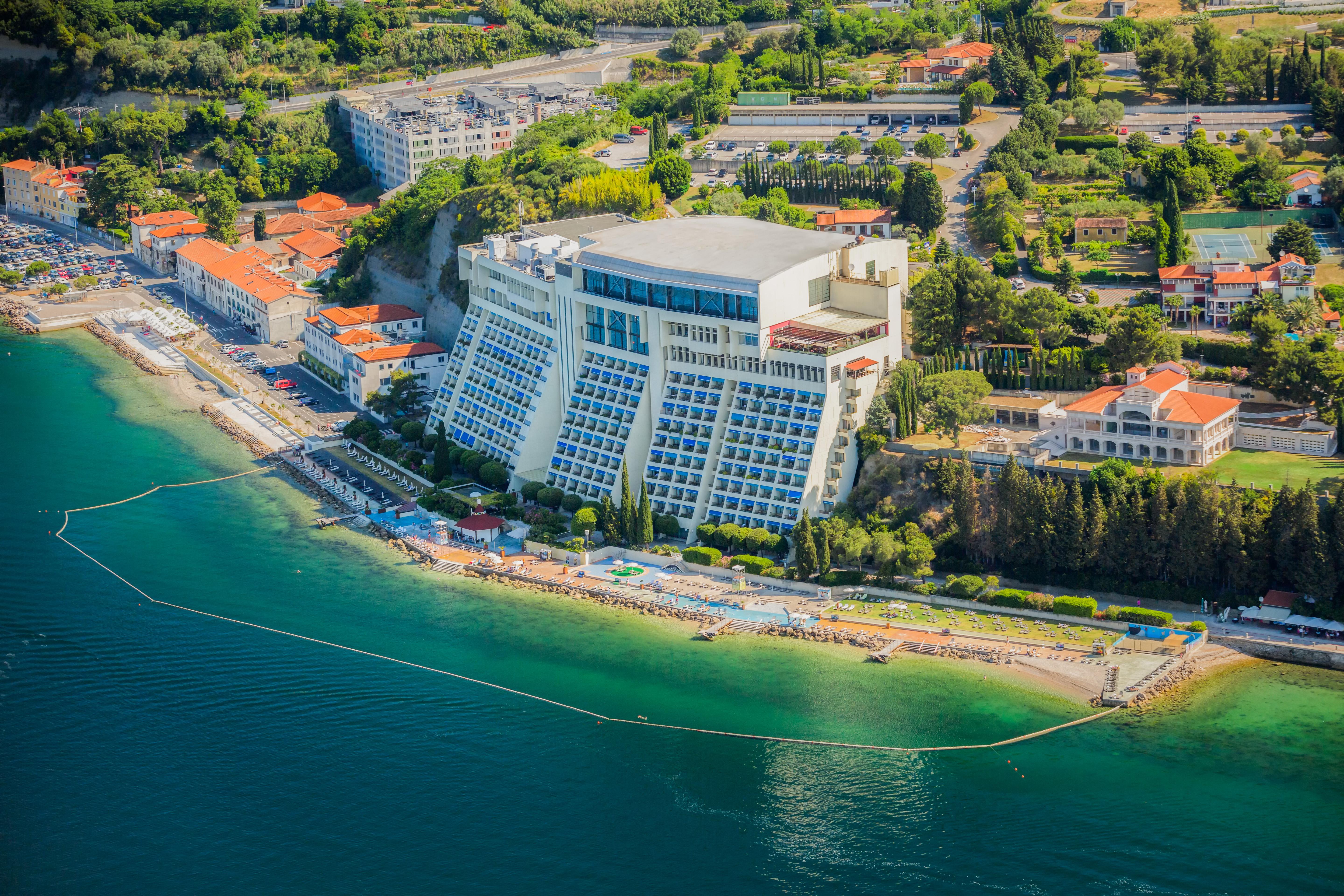 Grand Hotel Bernardin Portorose Esterno foto The photo shows an aerial view of a coastal area featuring a large hotel building situated on a beach. The hotel has multiple floors and appears to have a modern design, with a structured façade facing the water. Surrounding the hotel are various bui