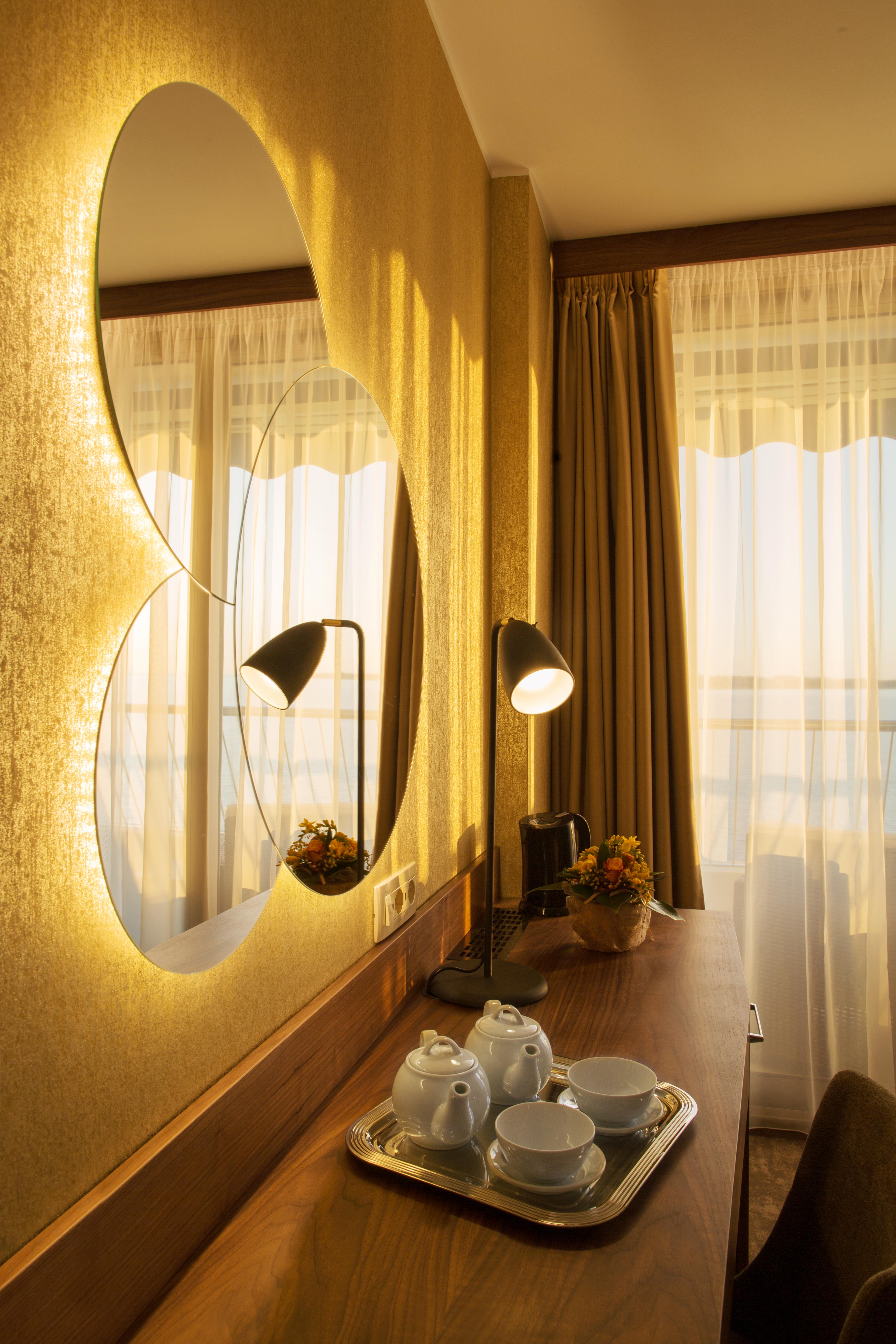 Grand Hotel Bernardin Portorose Esterno foto The photo features a cozy interior setting, likely from a hotel or a stylish room. There are two round mirrors on the wall, reflecting soft, warm light. A sleek black lamp sits on a wooden desk, providing additional illumination. Next to the lamp, th