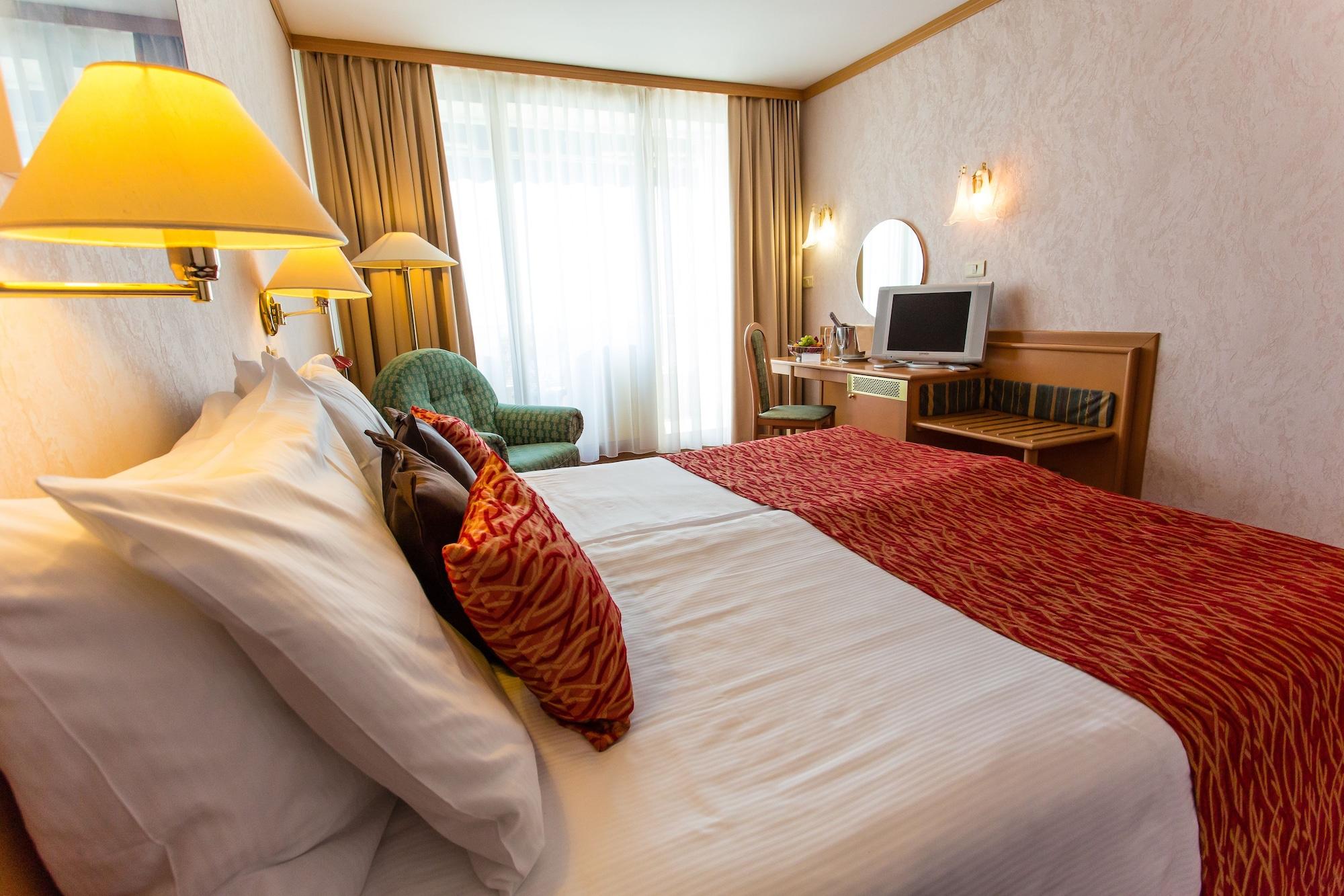 Grand Hotel Bernardin Portorose Esterno foto The photo shows a hotel room featuring two beds, each with decorative pillows. There is a warm color scheme, highlighted by a red bedspread. To the side, a green armchair is visible, along with a desk that has a mirror and a television on it. Large w