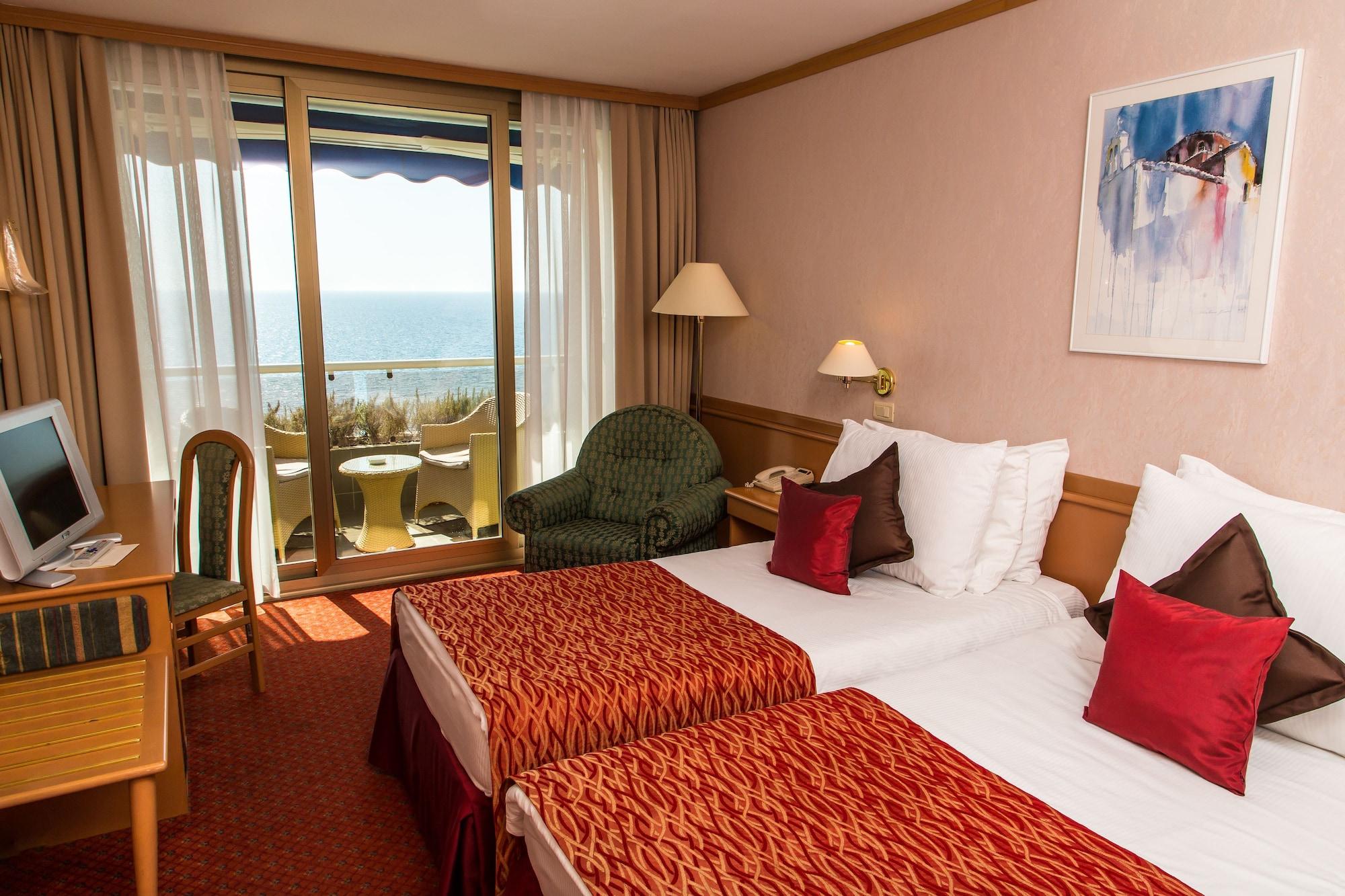 Grand Hotel Bernardin Portorose Esterno foto The photo shows a hotel room featuring two twin beds with decorative red and white bedding. There is a small seating area with a green chair and a side table. The room has large windows that offer a view of the ocean, and a balcony with two chairs is