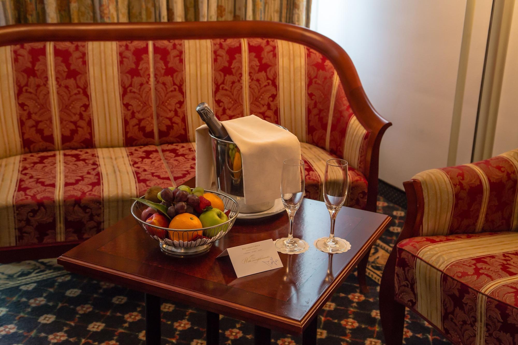 Grand Hotel Bernardin Portorose Esterno foto The photo depicts a cozy hotel room setting. In the center, there is a small wooden table adorned with a silver champagne bucket, a white cloth draped over it, and two champagne flutes. Next to it, there is a bowl of assorted fruits, including orange