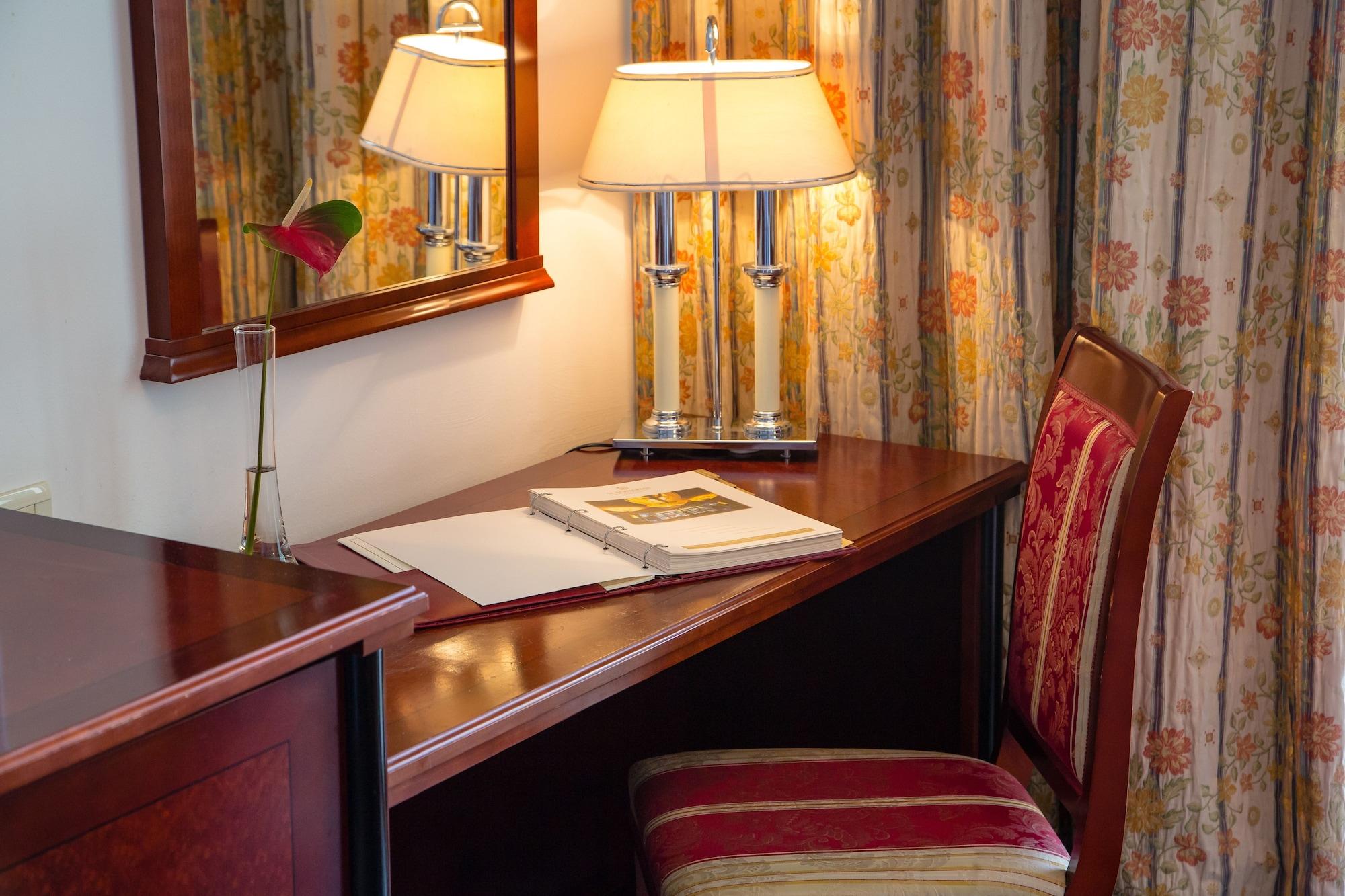 Grand Hotel Bernardin Portorose Esterno foto The photo shows a cozy desk setup in a room. There's a wooden desk with a dark finish next to a comfortable chair with a striped cushion. On the desk, a few items are placed, including an open book or notebook and a small vase with a flower. Above th
