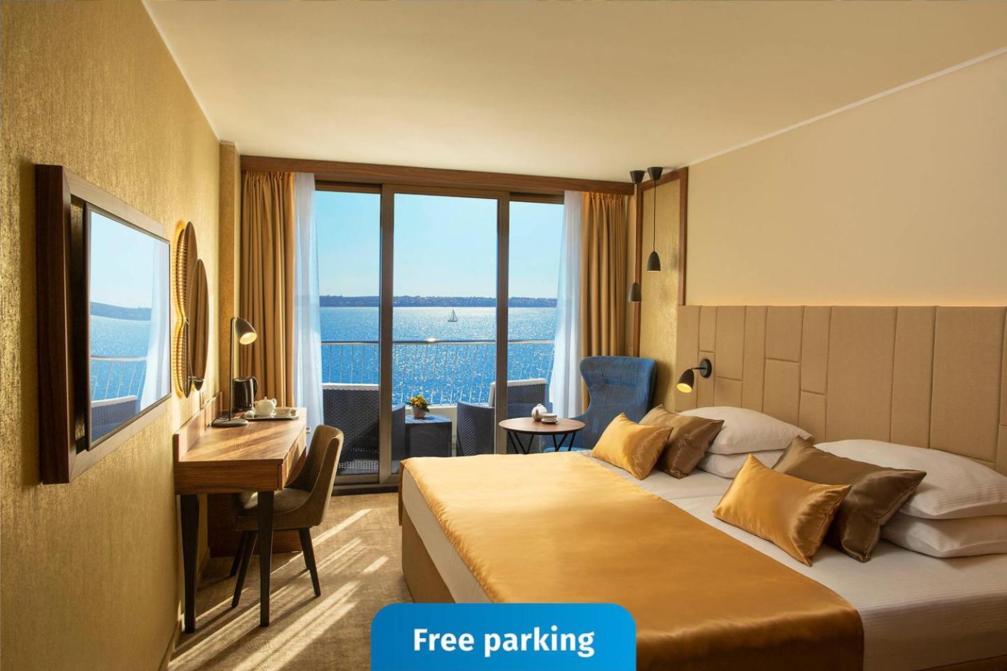 Grand Hotel Bernardin Portorose Esterno foto The photo shows a modern hotel room featuring a large window with a view of the water. The room is decorated in warm tones, with a comfortable bed that has gold-colored bedding. There is a desk and chair situated next to the window, as well as a wall