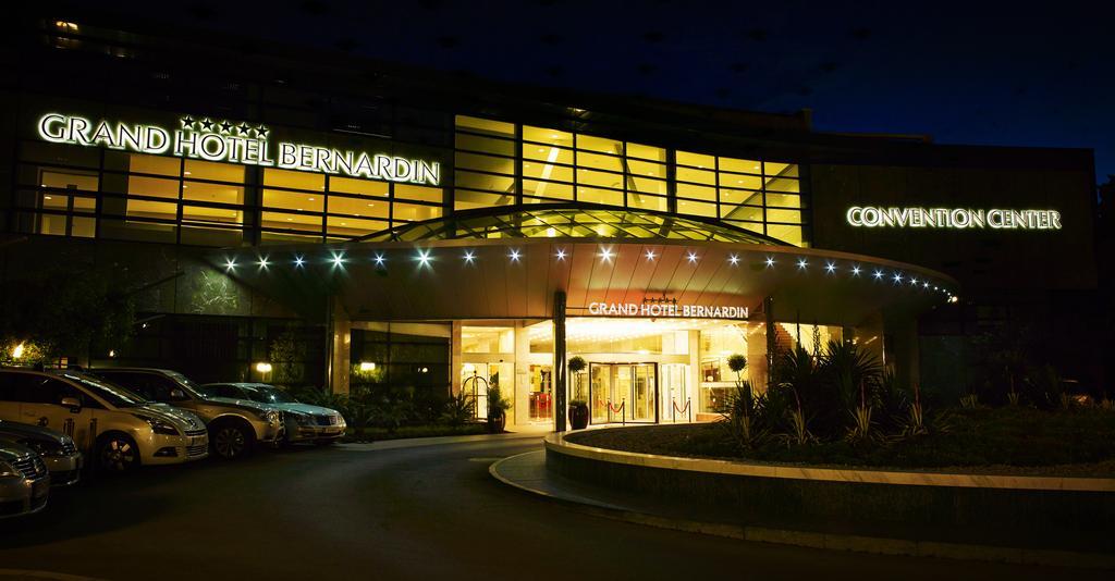 Grand Hotel Bernardin Portorose Esterno foto The photo shows a building, identified as the Grand Hotel Bernardin, illuminated at night. The entrance features a curved awning with decorative lights, creating a welcoming atmosphere. The front area has several parked cars and large glass windows, 