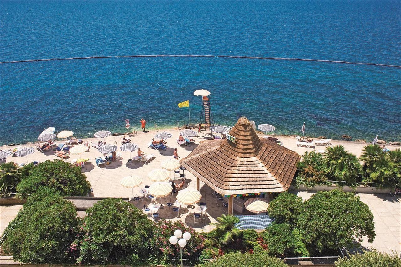 Grand Hotel Bernardin Portorose Esterno foto The photo shows a picturesque beach scene with clear blue water. There is a sandy area with several white umbrellas and lounge chairs where people are relaxing. A small thatched-roof gazebo or hut is visible, likely serving as a food or drink stand. 