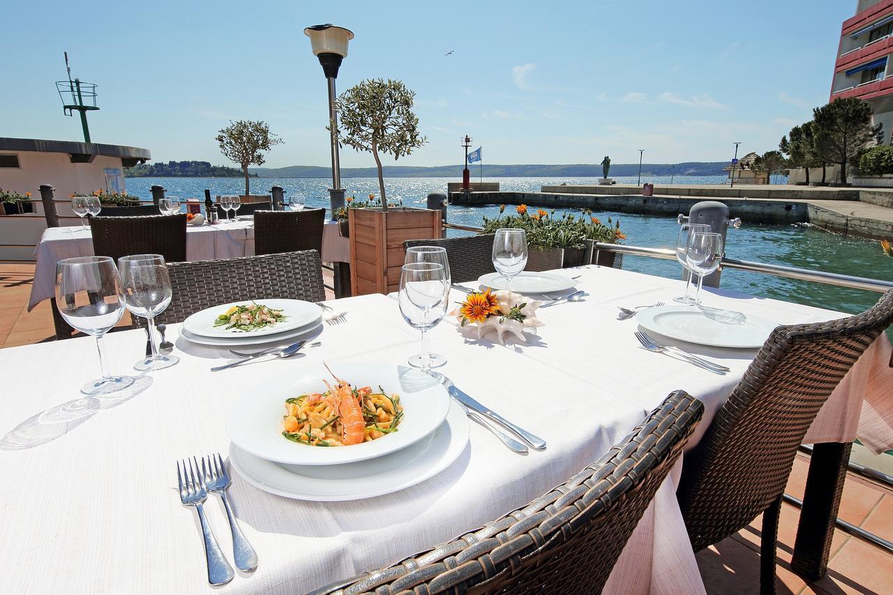 Grand Hotel Bernardin Portorose Esterno foto The photo shows a picturesque outdoor dining setup by a waterfront. There are several tables with elegant white tablecloths, each set with plates, cutlery, and wine glasses. In the foreground, a plate of pasta with salmon is prominently displayed alo