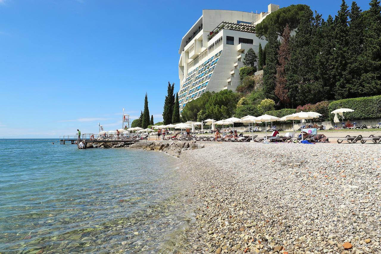Grand Hotel Bernardin Portorose Esterno foto The photo depicts a beach scene with a clear blue sky and calm waters. In the foreground, there is a pebbly shore where people are lounging, enjoying the sun, and some are swimming. Beach umbrellas provide shade for those relaxing on sunbeds. In the 