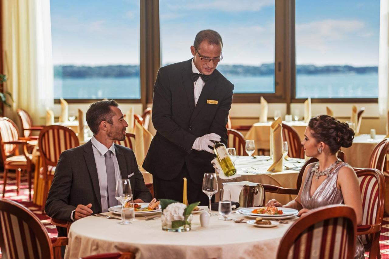 Grand Hotel Bernardin Portorose Esterno foto The image shows a fine dining setting in a restaurant. In the foreground, a well-dressed couple is seated at an elegantly set table. The man is looking towards the waiter, who is pouring a drink from a bottle into a wine glass. The woman is sitting b