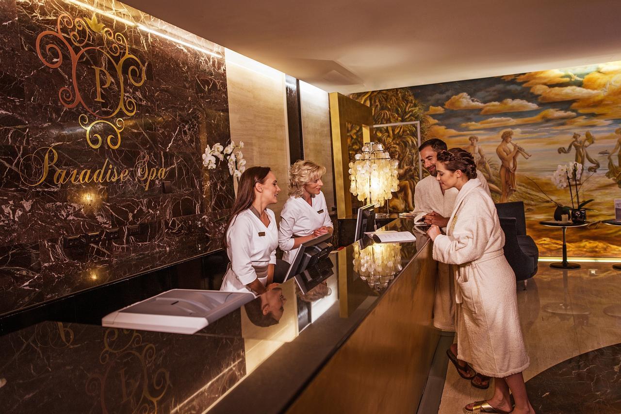 Grand Hotel Bernardin Portorose Esterno foto The photo shows a luxurious spa reception area. There are three staff members behind a sleek, dark marble reception desk, wearing white robes. Two guests, also in robes, are interacting with the staff. The background features an elegant mural and sty