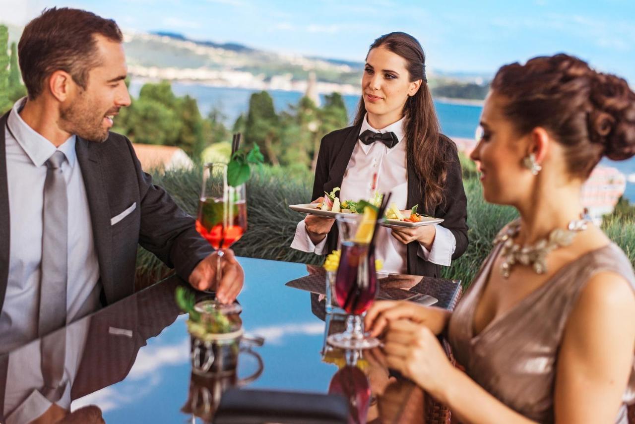 Grand Hotel Bernardin Portorose Esterno foto The photo shows a dining scene likely set in a restaurant with a picturesque view. In the image, a man and a woman are seated at a table, both dressed elegantly. The woman is wearing a sparkling dress, and the man is in a formal suit. A waitress, dre