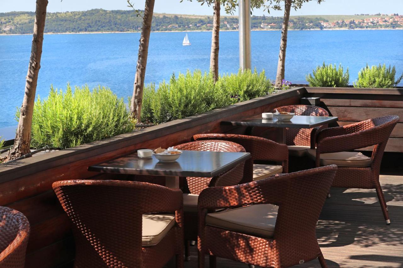 Grand Hotel Bernardin Portorose Esterno foto The photo shows an outdoor seating area of a restaurant or café with several wicker chairs and tables. The setting is surrounded by green plants and has a view of a calm body of water, likely a lake or sea, in the background. In the distance, there's