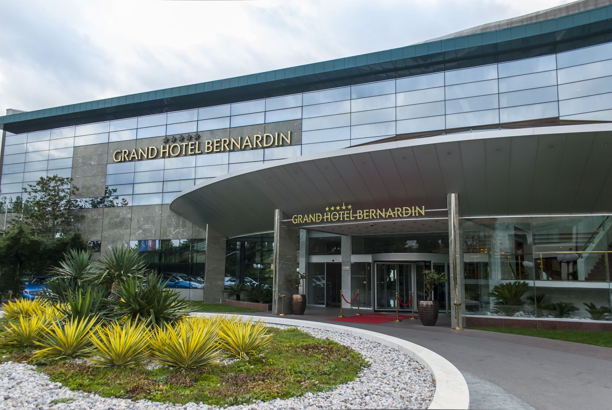 Grand Hotel Bernardin Portorose Esterno foto The photo showcases the entrance of the Grand Hotel Bernardin, featuring a modern architectural design. The facade includes large glass windows and a sleek overhang. Strategically placed greenery, such as palms and low shrubs, adds to the aesthetic a